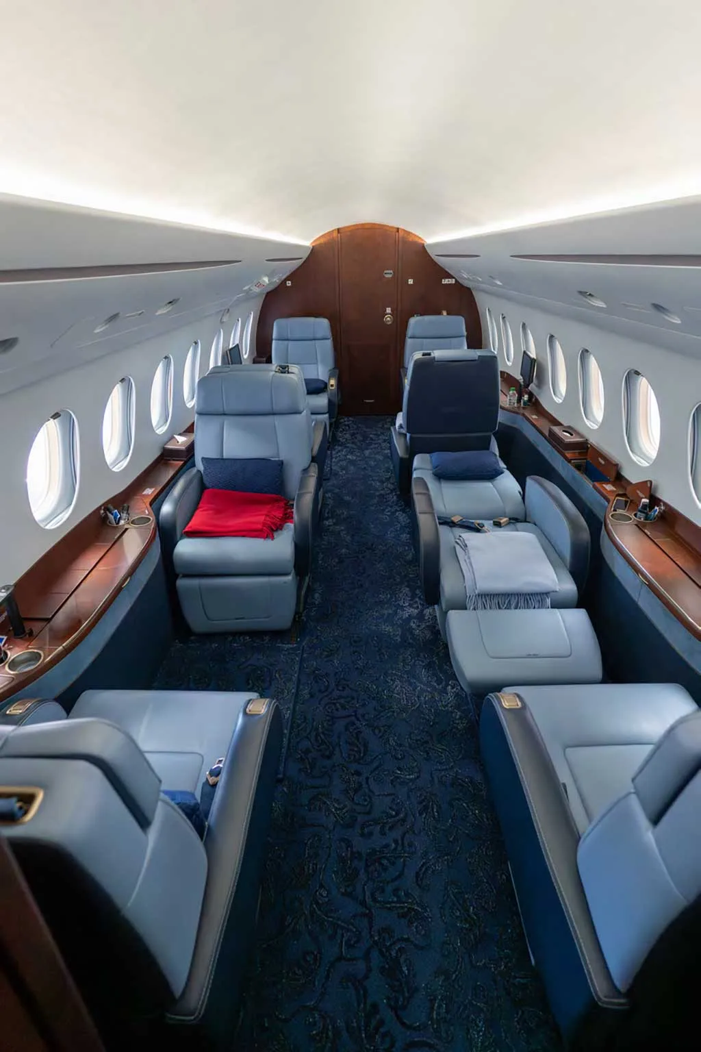 A long view of the business jet's cabin, featuring multiple rows of light blue leather seats, dark blue patterned carpet, and polished wood trim.