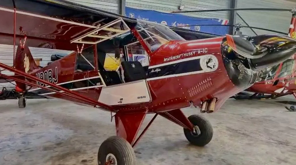 A front-side view of the burgundy Aviat Aircraft Husky A-1C, registration N90BJ, with its door open, parked in a hangar.