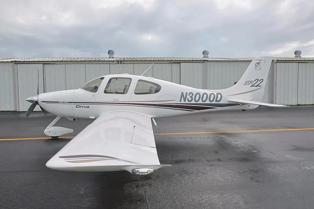 Left side view of a white Cirrus SR22 aircraft with N3000D registration parked on a wet tarmac next to a hangar.