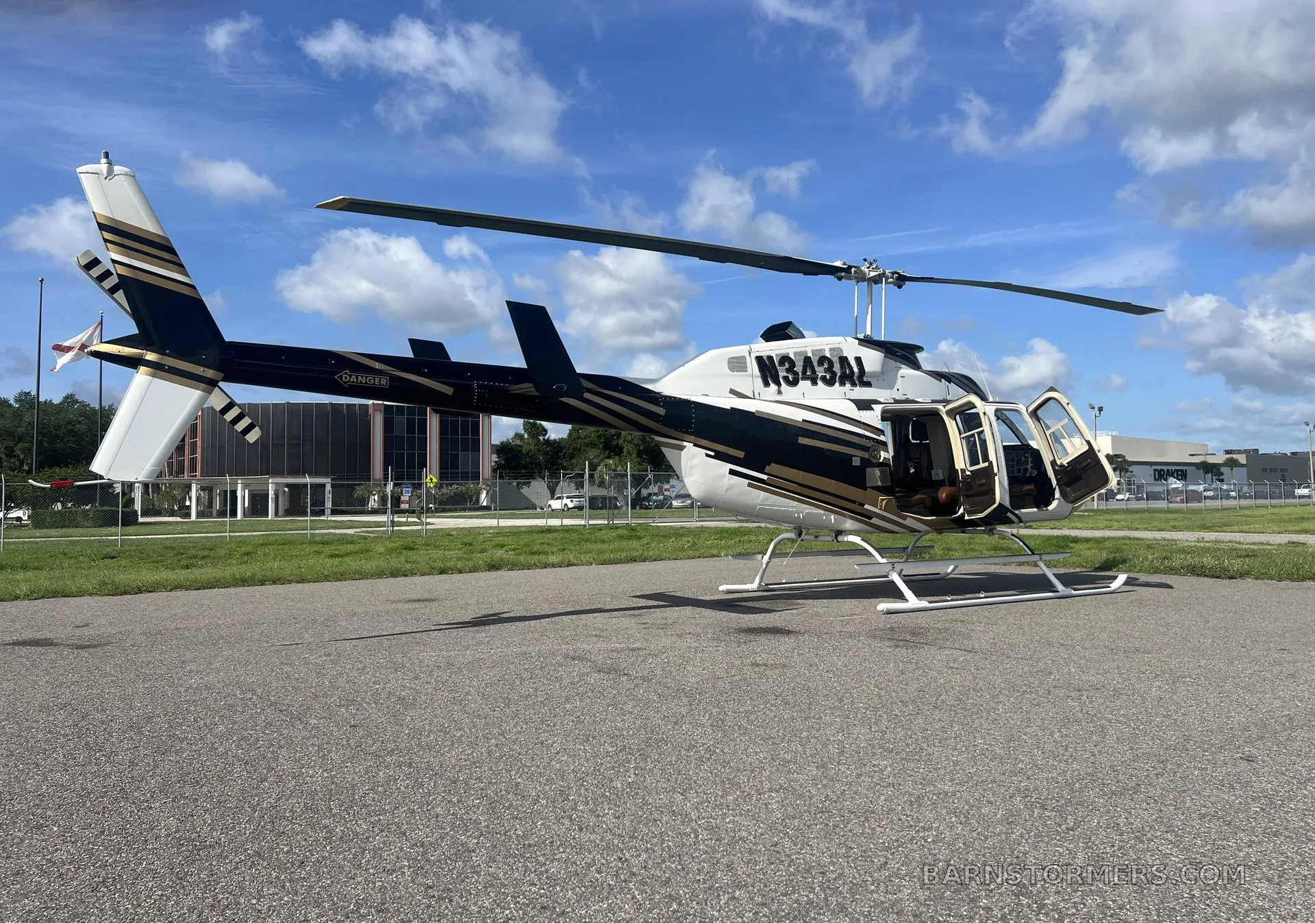 A white and gold Bell 206 helicopter (N343AL) with its doors open is parked on an asphalt pad under a blue sky.