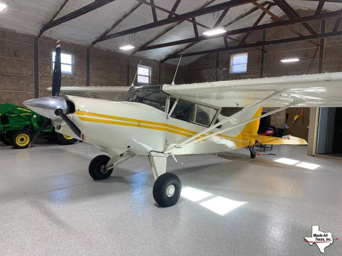 A white single-engine propeller aircraft with yellow stripes is parked in a hangar, viewed from the front-left.