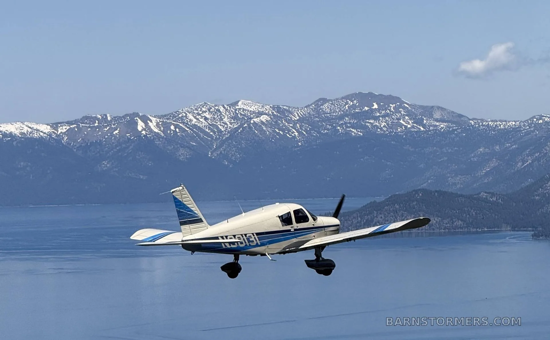 A white and blue single-engine propeller aircraft (N9813I) flies low over a lake with snow-capped mountains in the background.