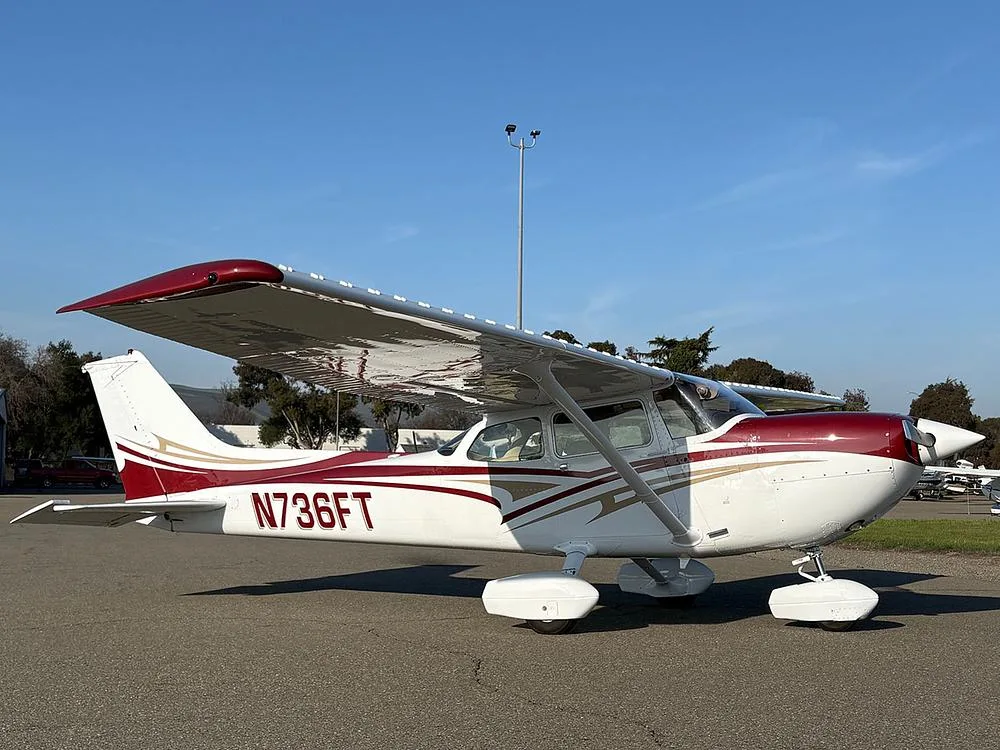 A white and maroon Cessna 172 with gold accents, parked on an airfield with trees and hills in the background.