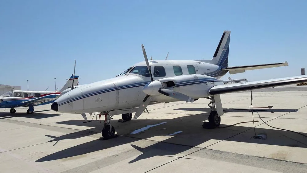 A white Piper PA-31-350 Navajo Chieftain with blue stripes is parked on an airport tarmac, viewed from the front-right.