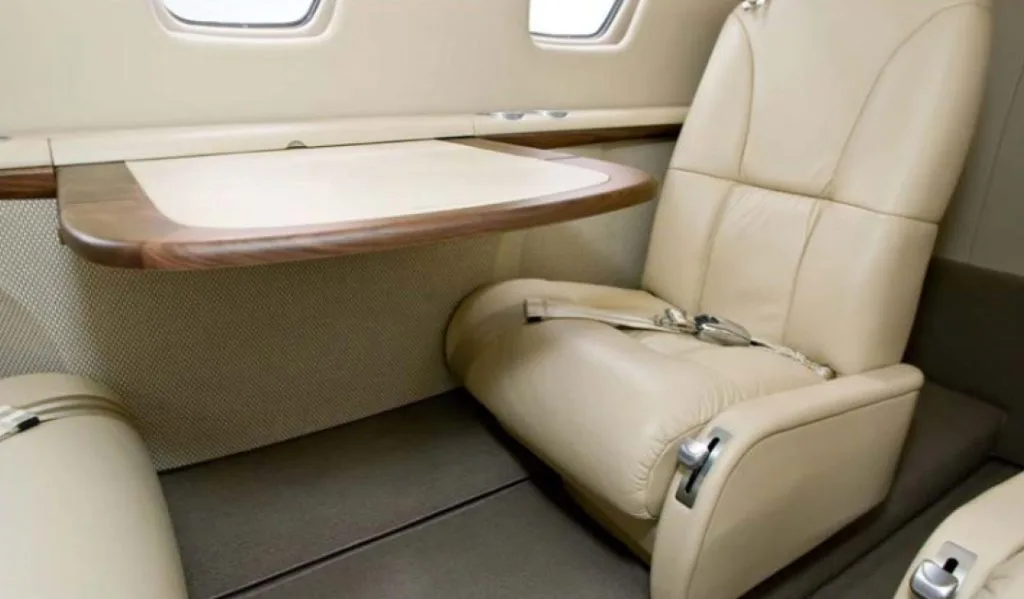 A close-up view of a beige leather seat and a wooden fold-out table in an aircraft cabin.