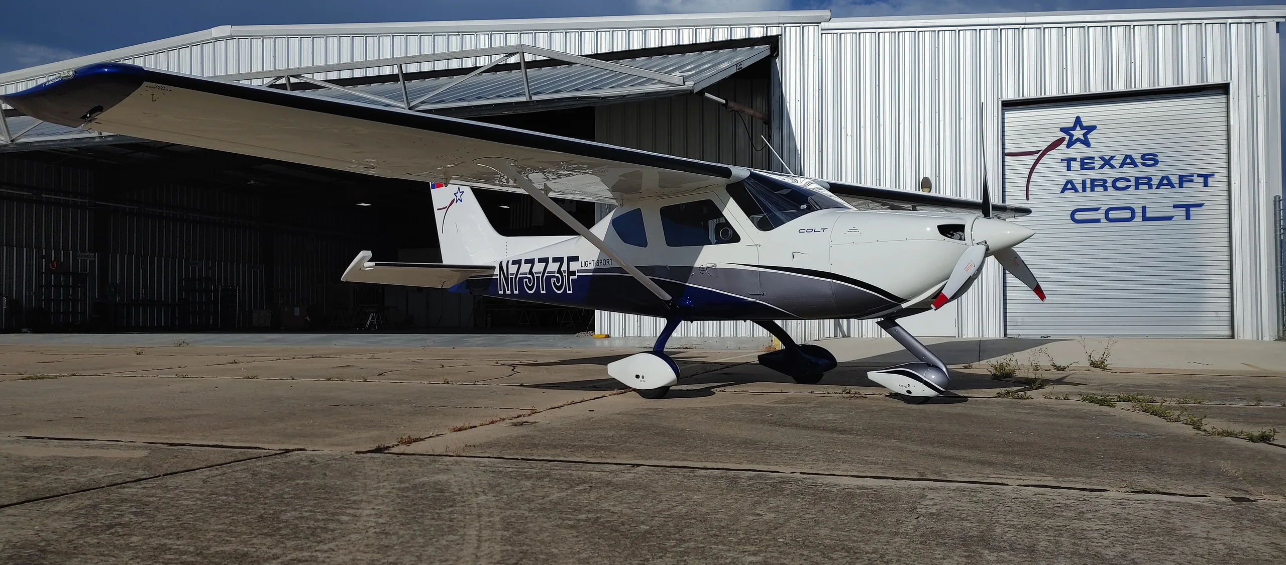 A white and blue Texas Aircraft Colt N7373F parked on a tarmac in front of a hangar.