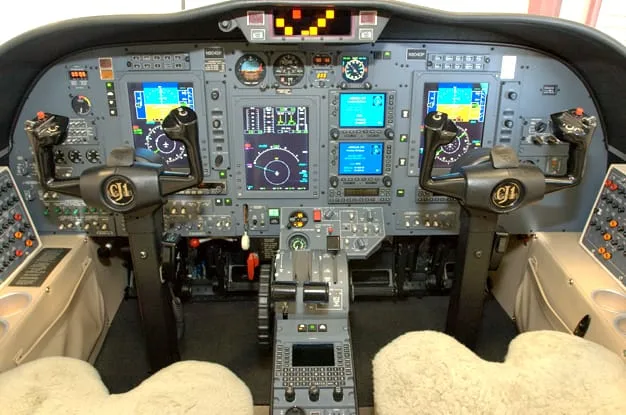 The cockpit of a modern business jet, featuring multiple digital displays, control yokes, and a central throttle quadrant.