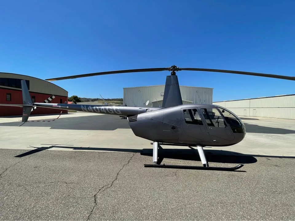 A grey Robinson R44 helicopter parked on an airport tarmac with hangars in the background under a clear blue sky.