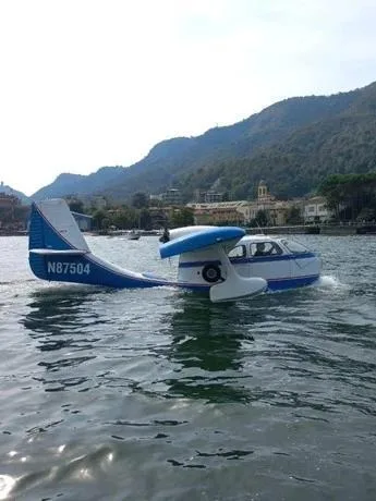 A blue and white amphibious aircraft, N87504, on the water with mountains and buildings in the background.
