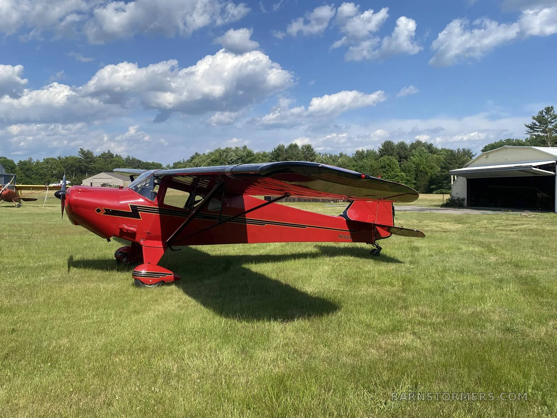 A red single-engine high-wing aircraft with black wings and stripes, parked on a grassy airfield under a blue sky with clouds.