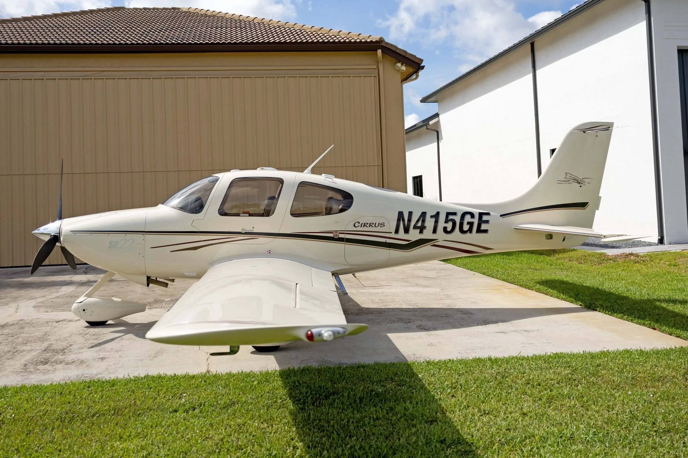 A white Cirrus SR22 aircraft with black and maroon stripes, registration N415GE, parked on a concrete pad next to a tan building.