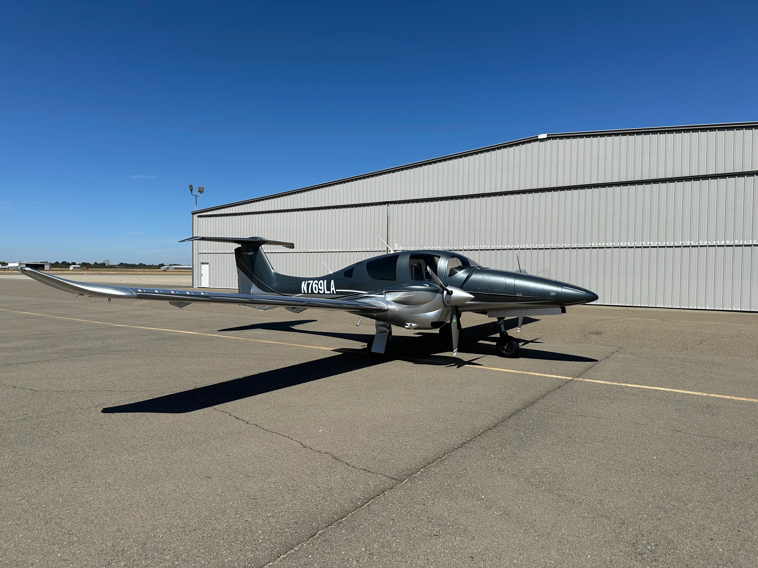 A grey and silver Diamond DA62 aircraft with registration N769LA parked on an airfield next to a hangar.