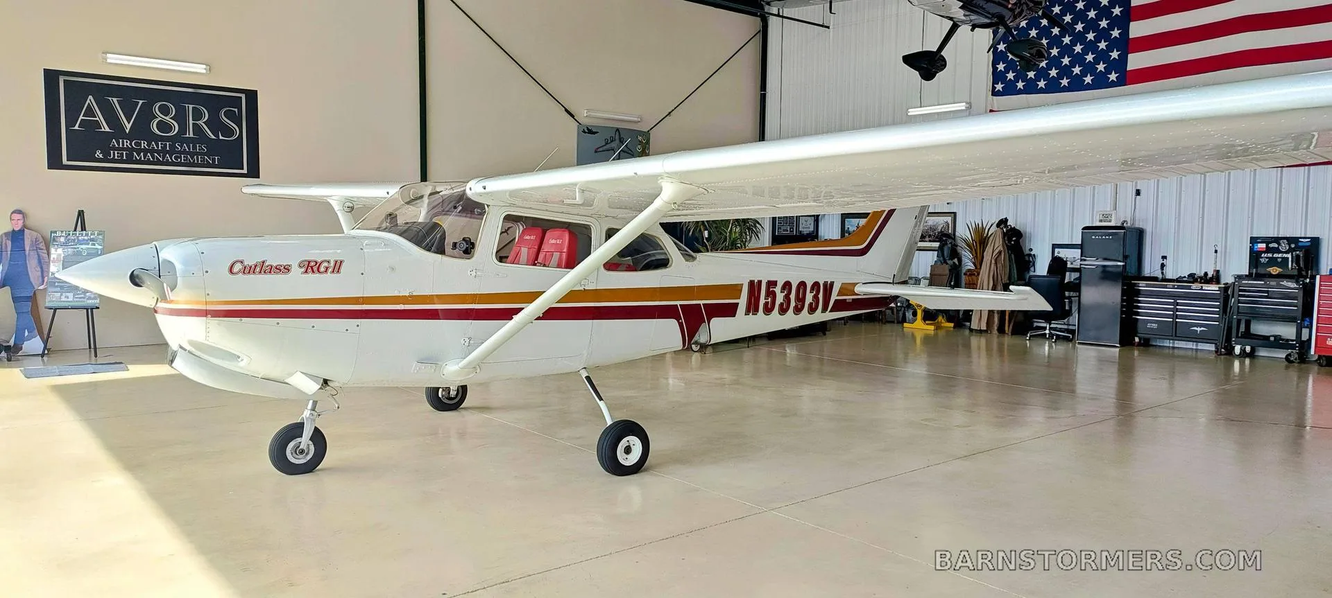 A white Cessna Cutlass RGII (N5393V) with red and gold stripes is parked inside a hangar with an American flag.