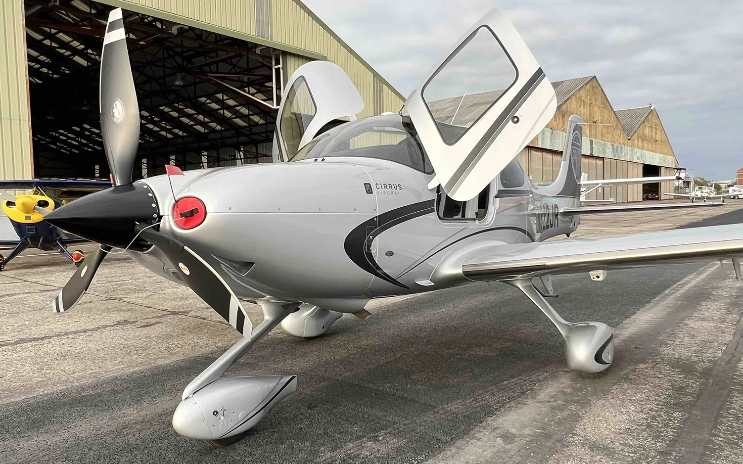 A silver Cirrus SR22 aircraft with black accents and open doors is parked in front of a hangar.