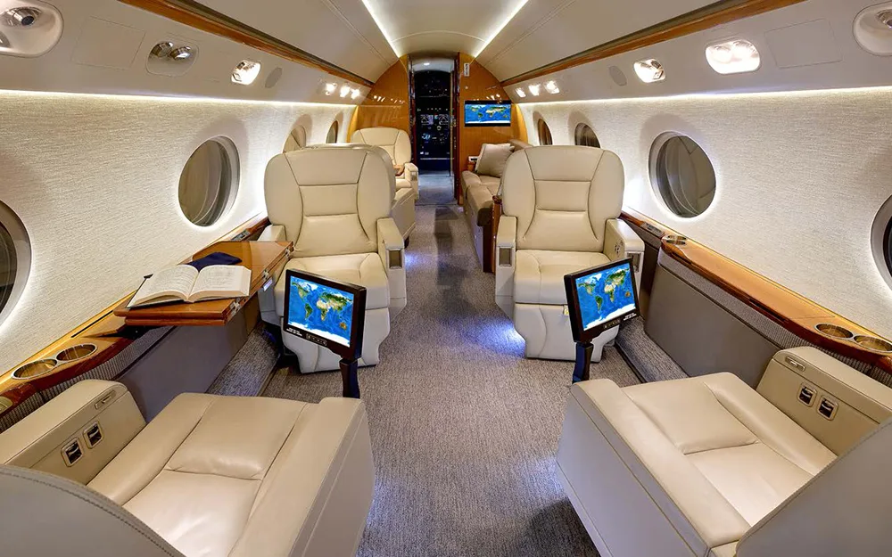 The aircraft cabin with multiple beige seats, individual entertainment screens displaying a world map, and a book on a side table.