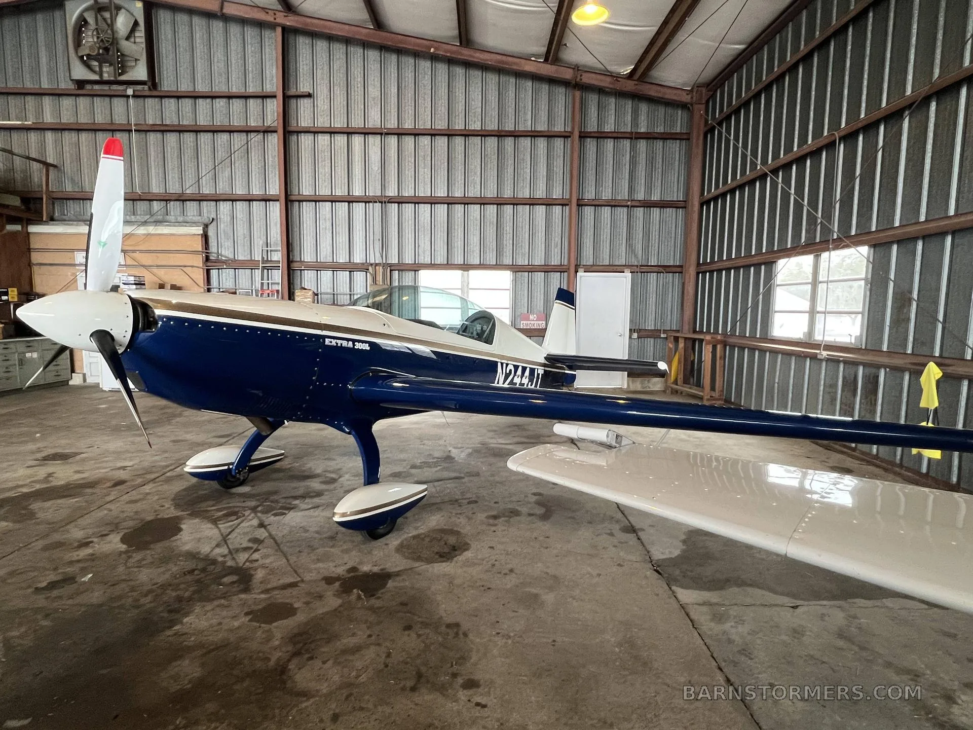 A blue and white Extra 300L aircraft with tail number N244JT is parked inside a metal hangar, viewed from the front-left.