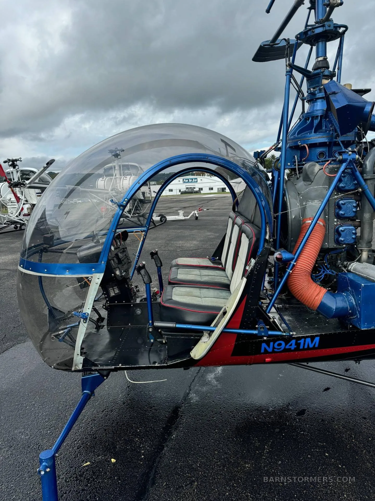 A side view of a blue and black helicopter on a wet tarmac, showing the clear bubble cockpit and exposed engine.