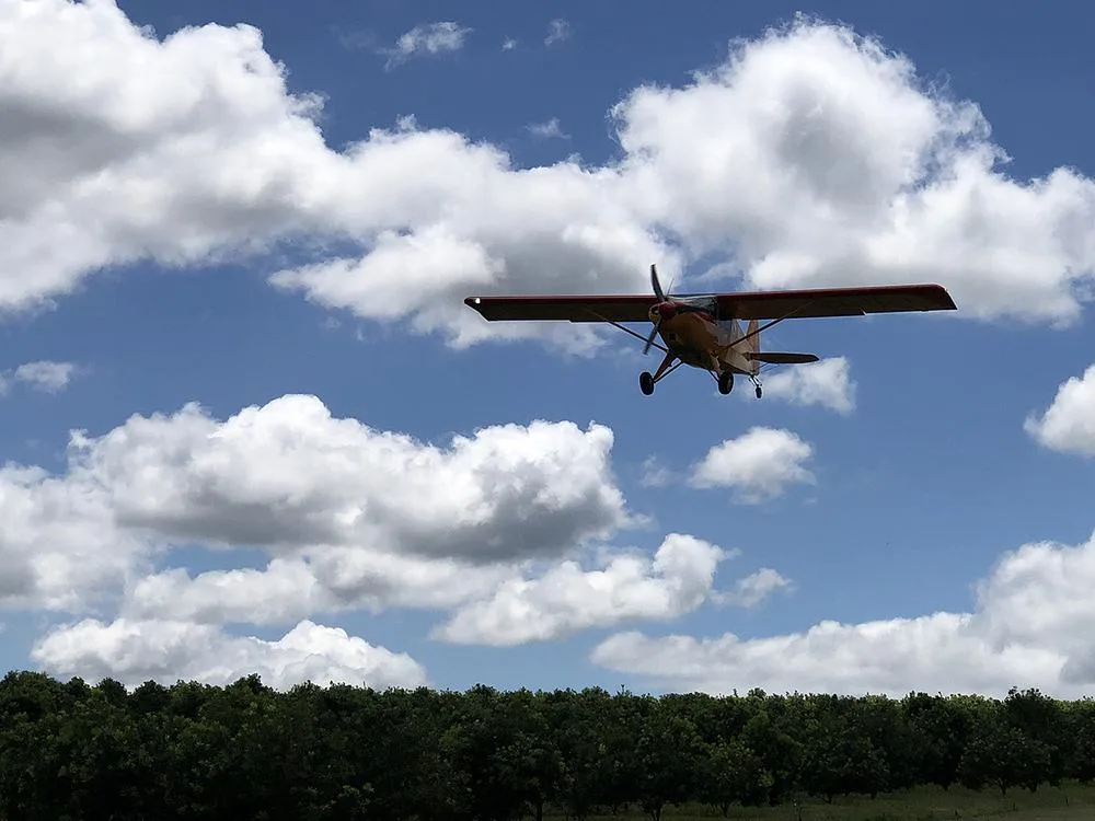 A red and orange Bearhawk Bravo aircraft flies low over a line of green trees under a partly cloudy blue sky.