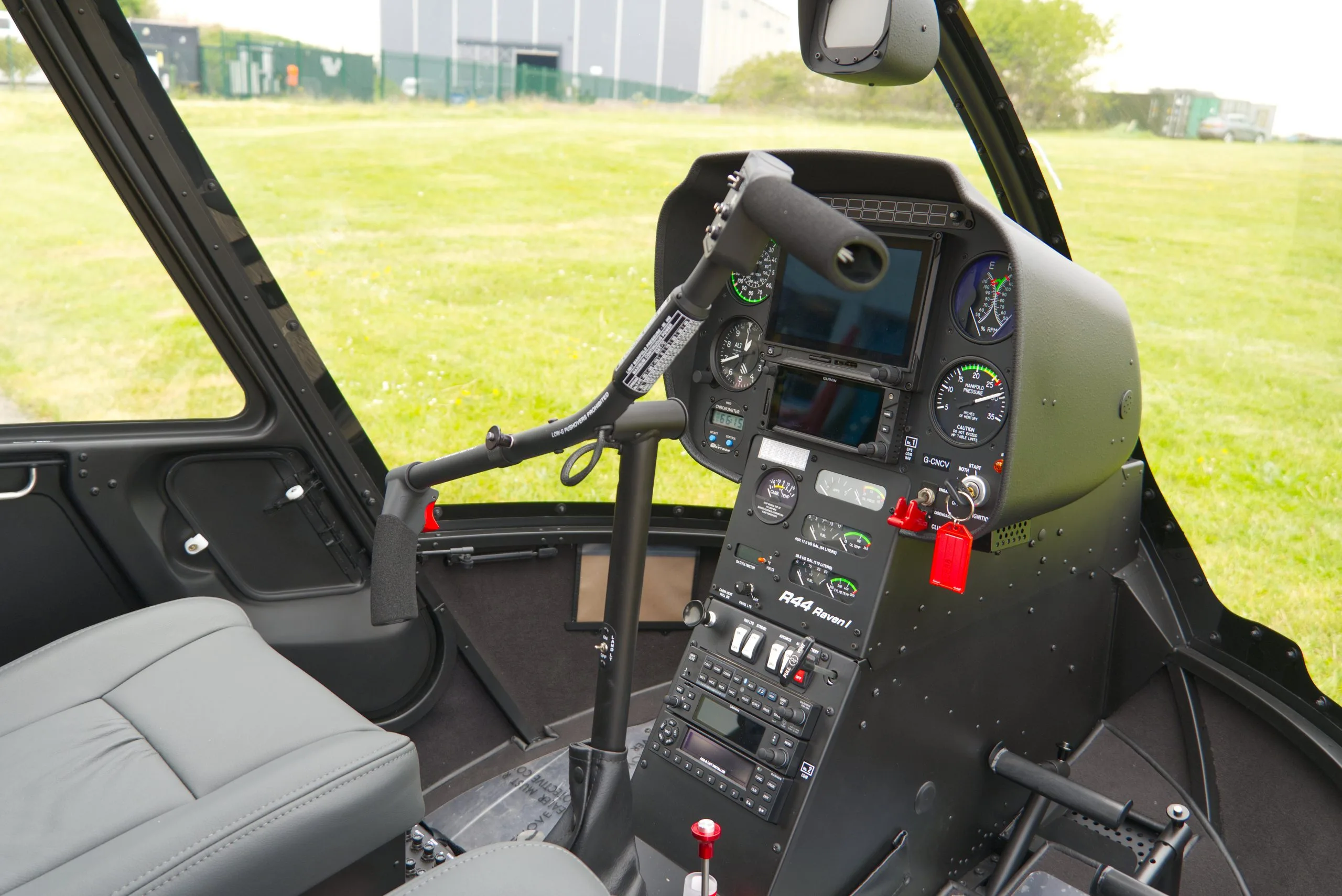 The black instrument panel and controls of a Robinson R44 Raven I helicopter cockpit, with gray seats and a view of a grassy field outside.