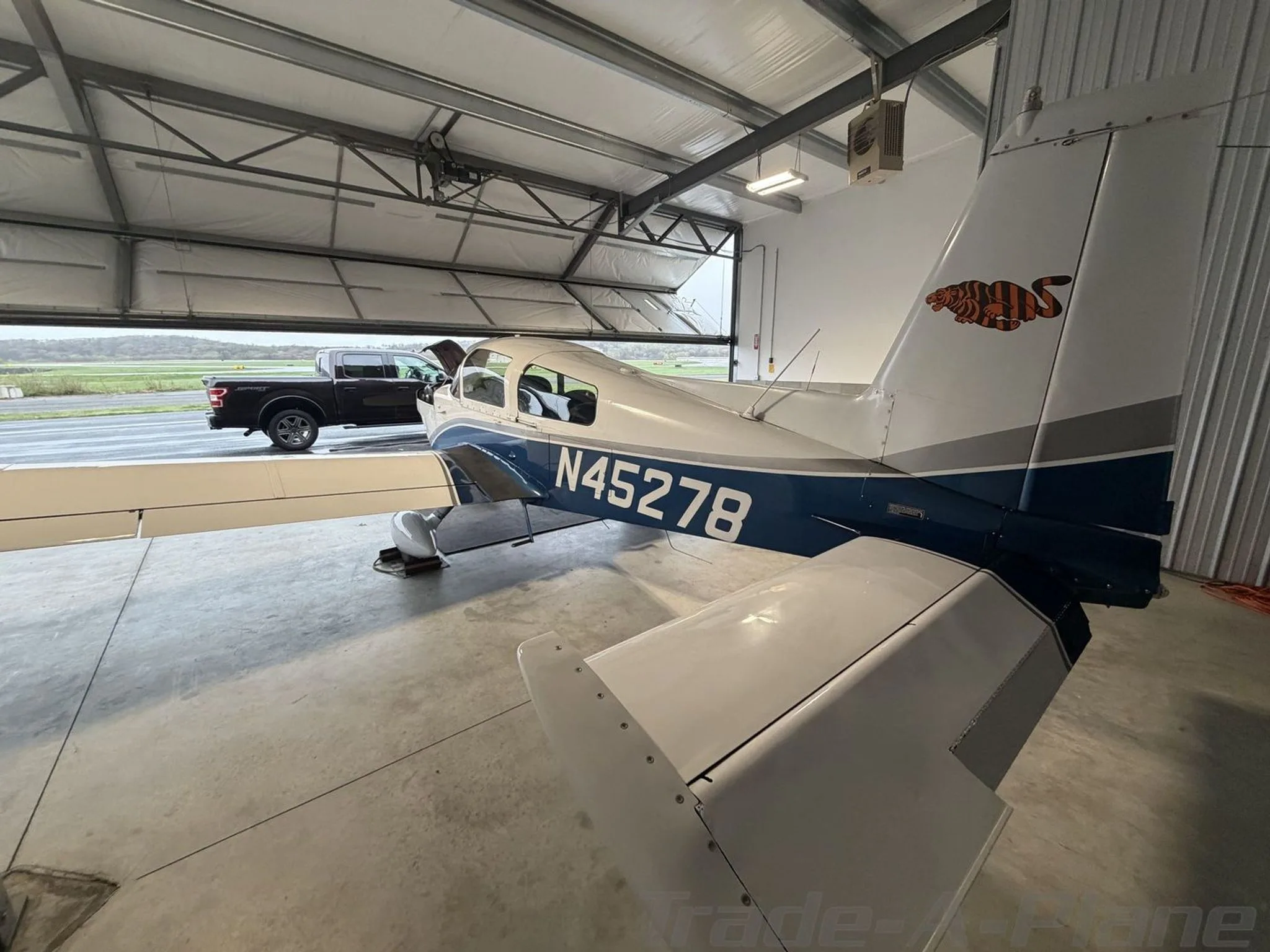 Rear-right view of a white and blue Grumman AA-5B Tiger N45278 in a hangar, with a truck visible outside.