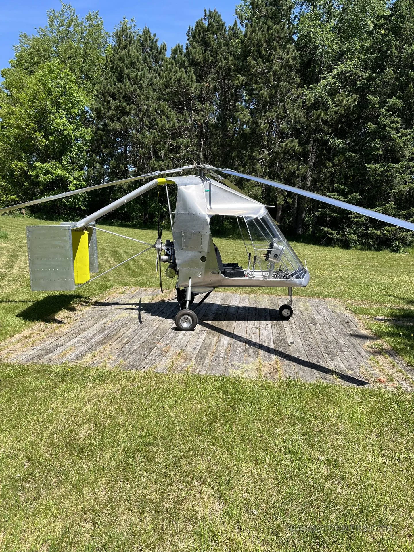 A silver single-seat ultralight helicopter/autogyro with a yellow tail fin, viewed from the side, parked on a wooden pad in a grassy field.