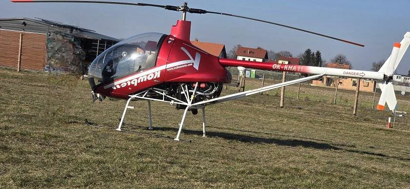 A red and white Kompress helicopter with registration OK-KHA11 is parked on a dry grassy field with buildings in the background.