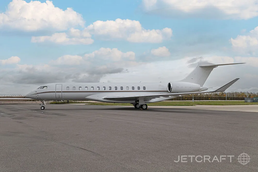 A full side view of a white Bombardier Global business jet with brown accent stripes parked on an airfield.