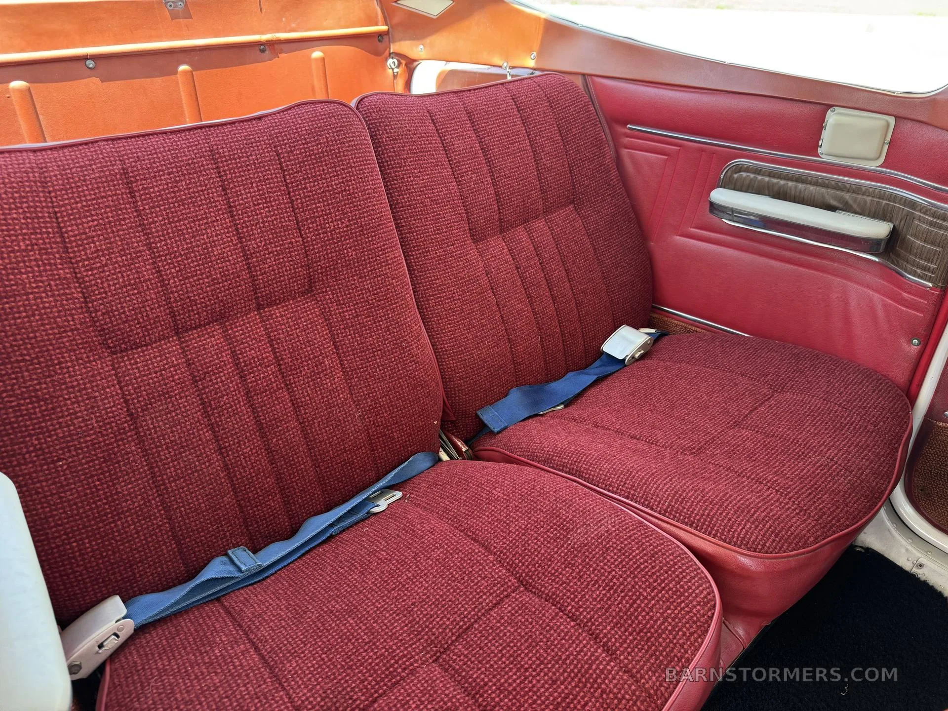 Two red fabric rear passenger seats with blue seatbelts in a small aircraft cabin.