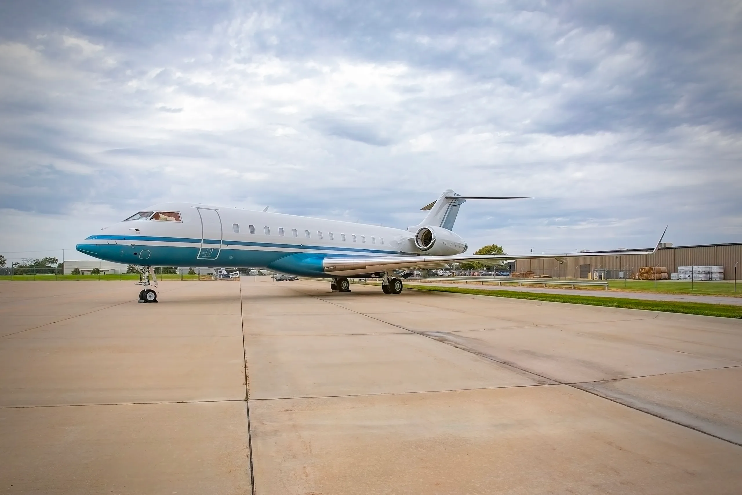A white and blue business jet, likely a Bombardier Global, parked on an airport tarmac under a cloudy sky.