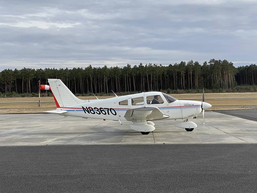 A white Piper Archer II aircraft with red and blue stripes on a tarmac, with a forest in the background.