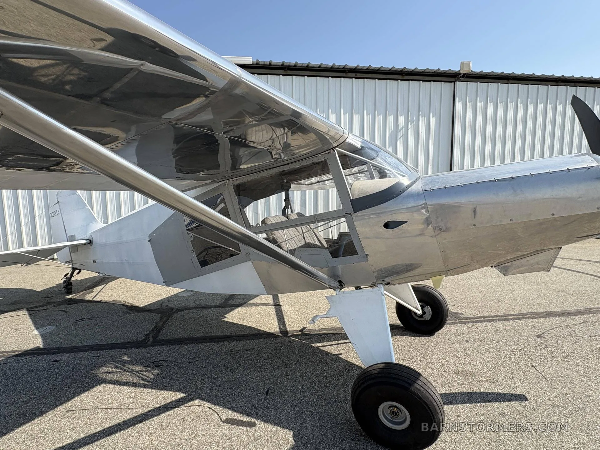 A side view of a silver and white single-engine aircraft, registration N213TJ, parked on an asphalt tarmac next to a white hangar.