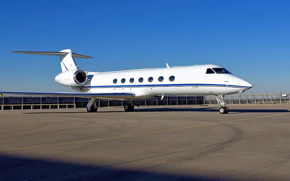 A white Gulfstream business jet with a blue stripe on the tarmac under a clear blue sky.