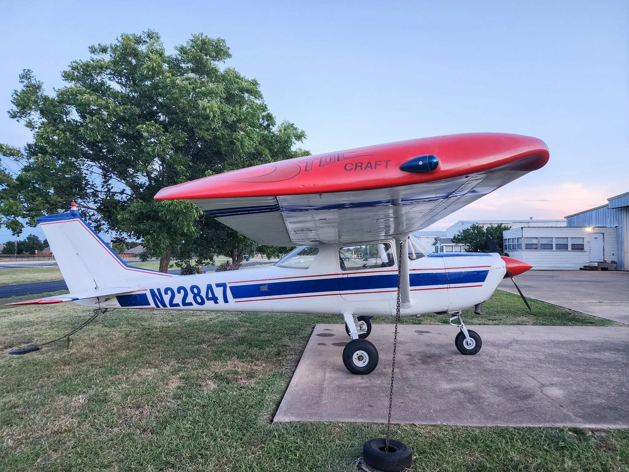 A white Cessna 150 with blue stripes and a red Horton STOL kit on the wing, parked on a concrete pad at dusk.