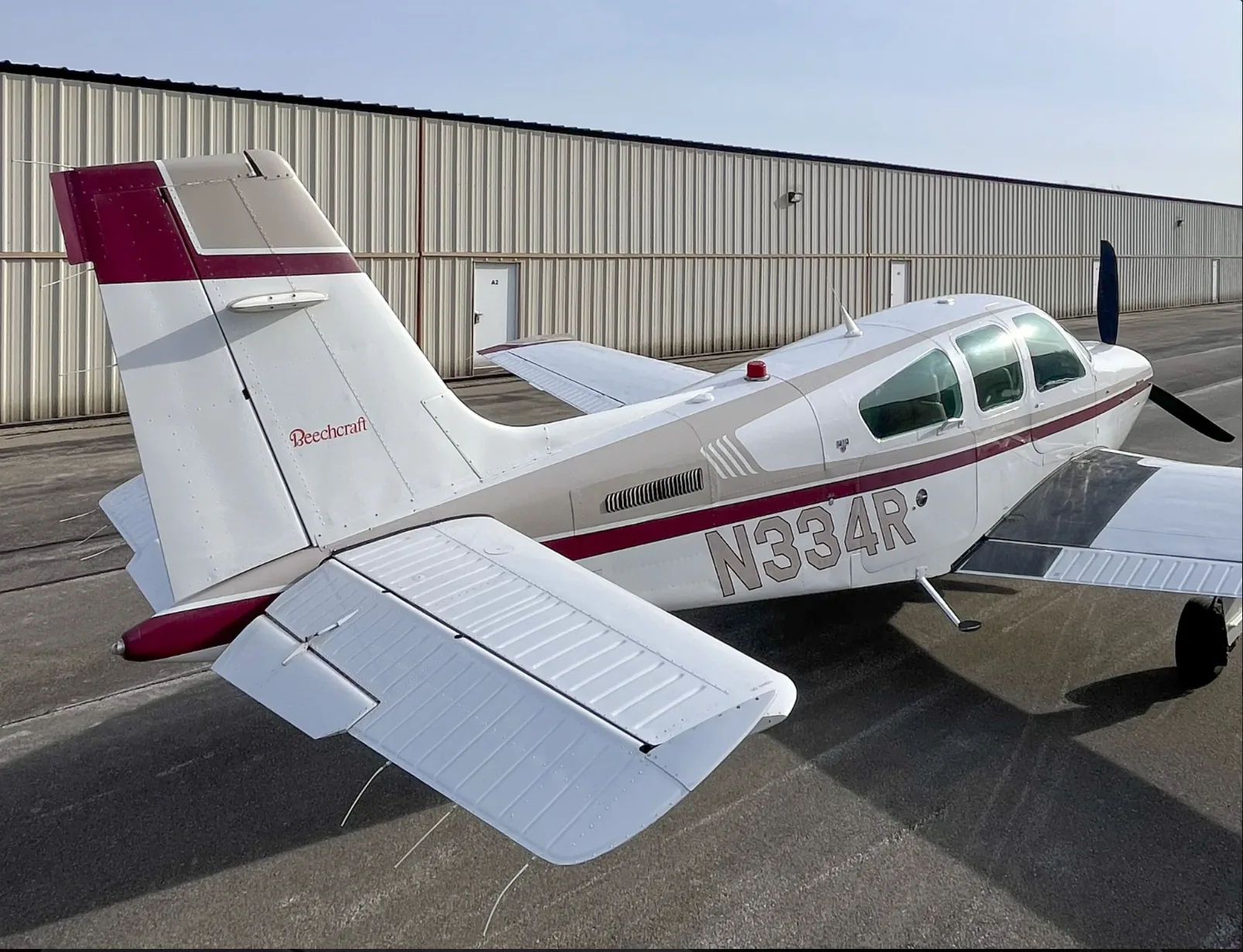 A rear-quarter view of a white and maroon Beechcraft Bonanza aircraft, tail number N334R, parked next to a hangar.