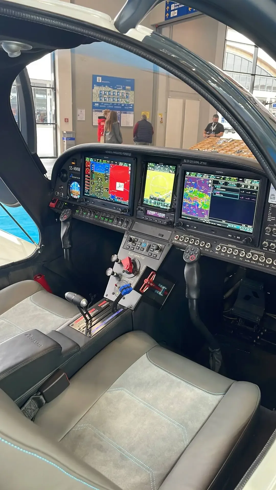 The interior of a light aircraft cockpit and cabin, showing grey seats, control sticks, and multiple display screens.