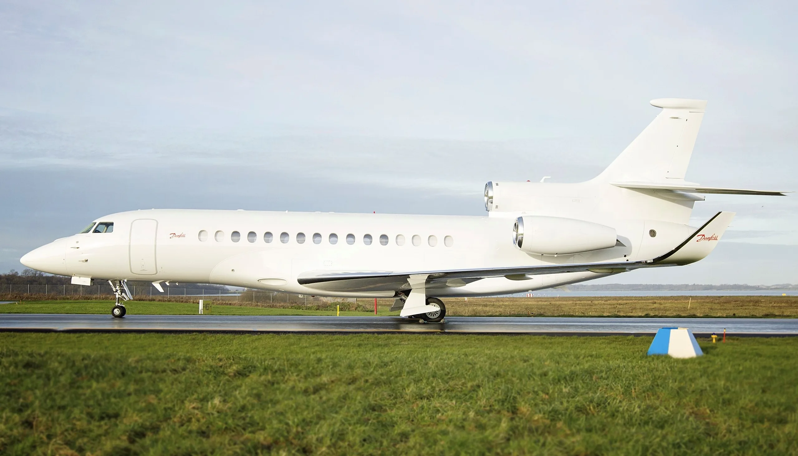 A white Dassault Falcon business jet with 'Danfoss' branding is parked on a tarmac with green grass in the foreground.