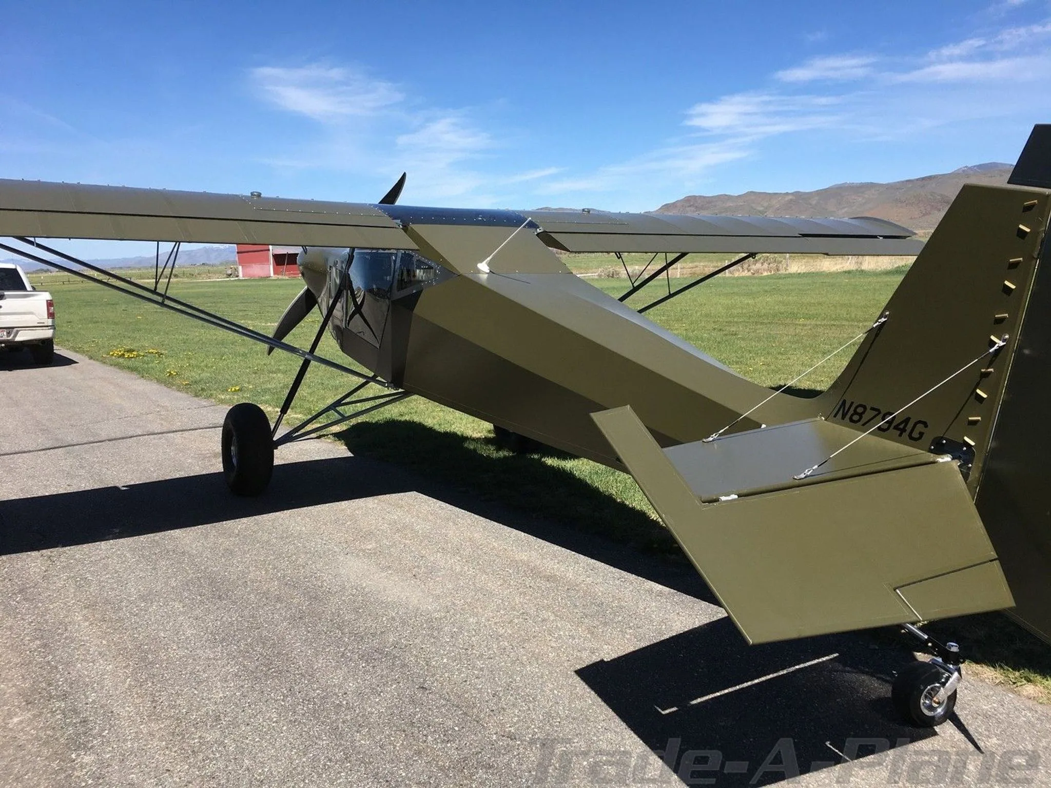 Rear quarter view of the green experimental aircraft N8794G on an airfield with a red building in the distance.