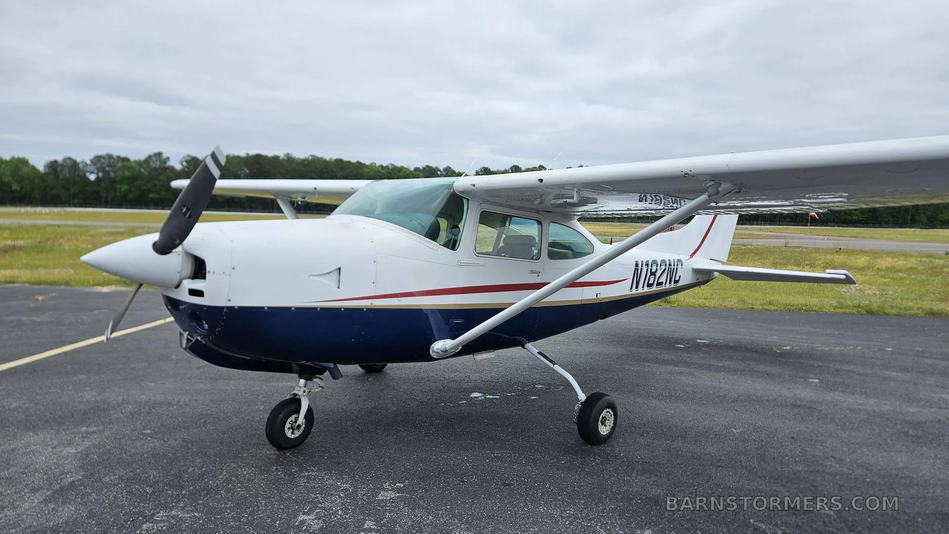 A white and blue Cessna 182 aircraft with red and gold stripes, parked on an asphalt tarmac under a cloudy sky.
