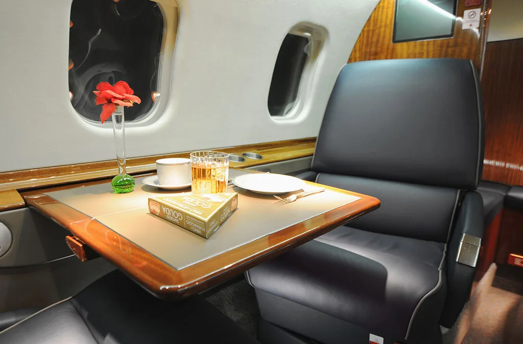 A private jet cabin table is set with a drink, a cup, a plate, and a red flower in a vase next to a black leather seat.