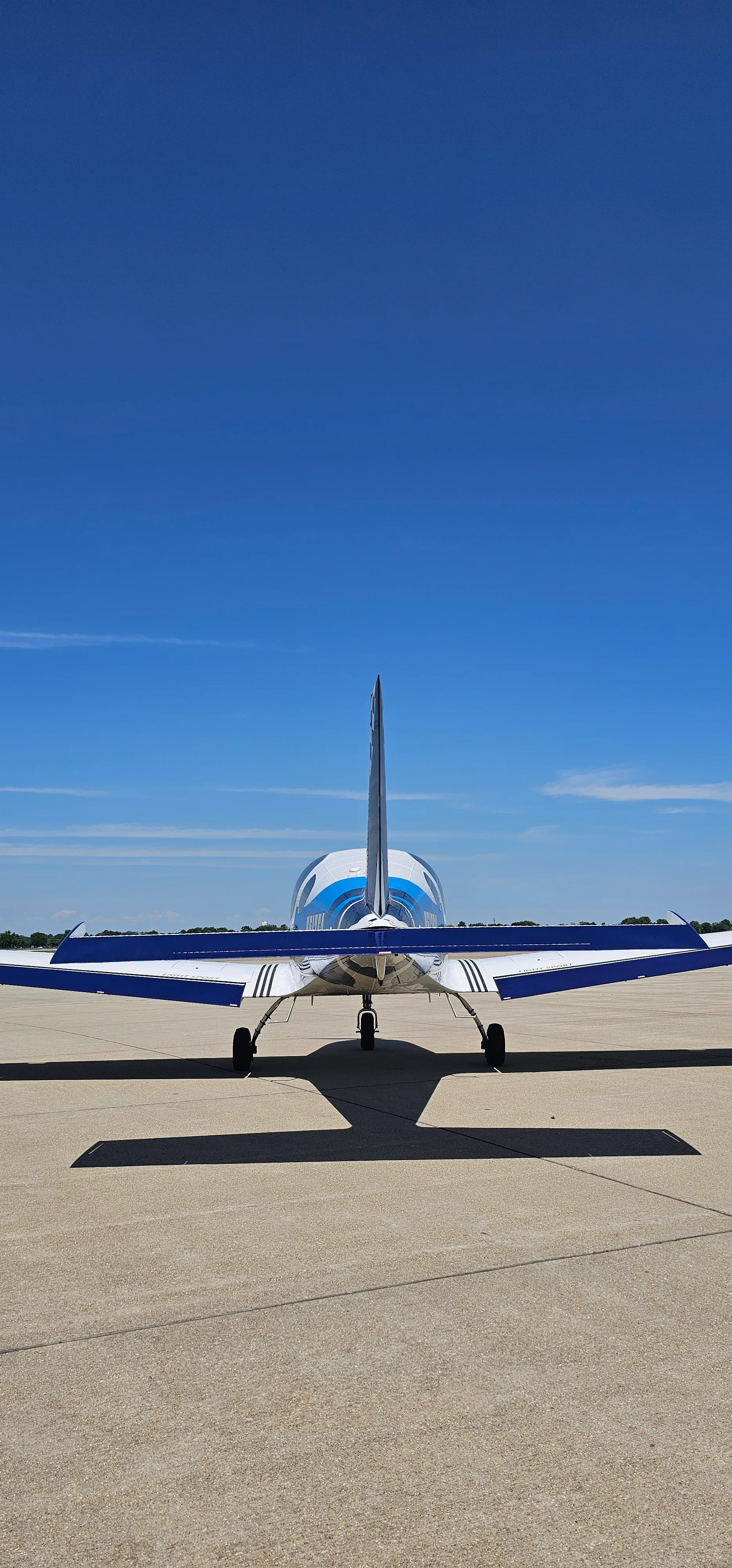 A rear view of a white and blue light sport aircraft with a T-tail, parked on an airfield under a clear blue sky.