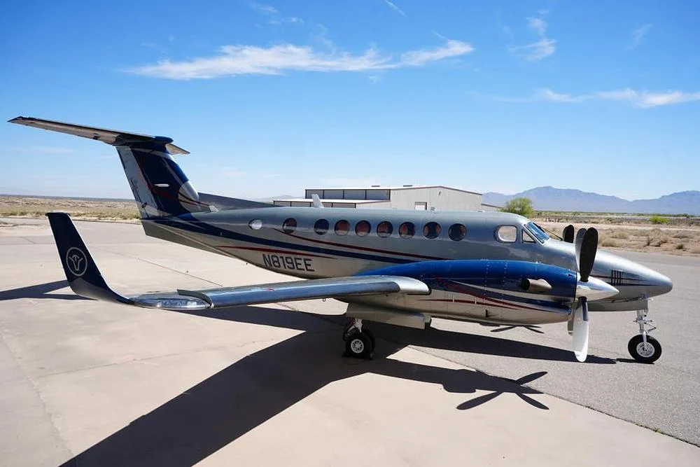 A side view of the silver and blue twin-turboprop aircraft N819EE, parked on an airfield under a blue sky.