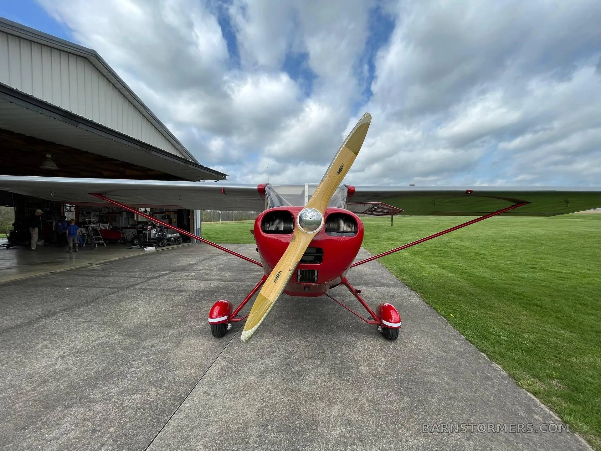 A head-on view of a red single-engine propeller aircraft with a wooden propeller, parked on concrete in front of a hangar.