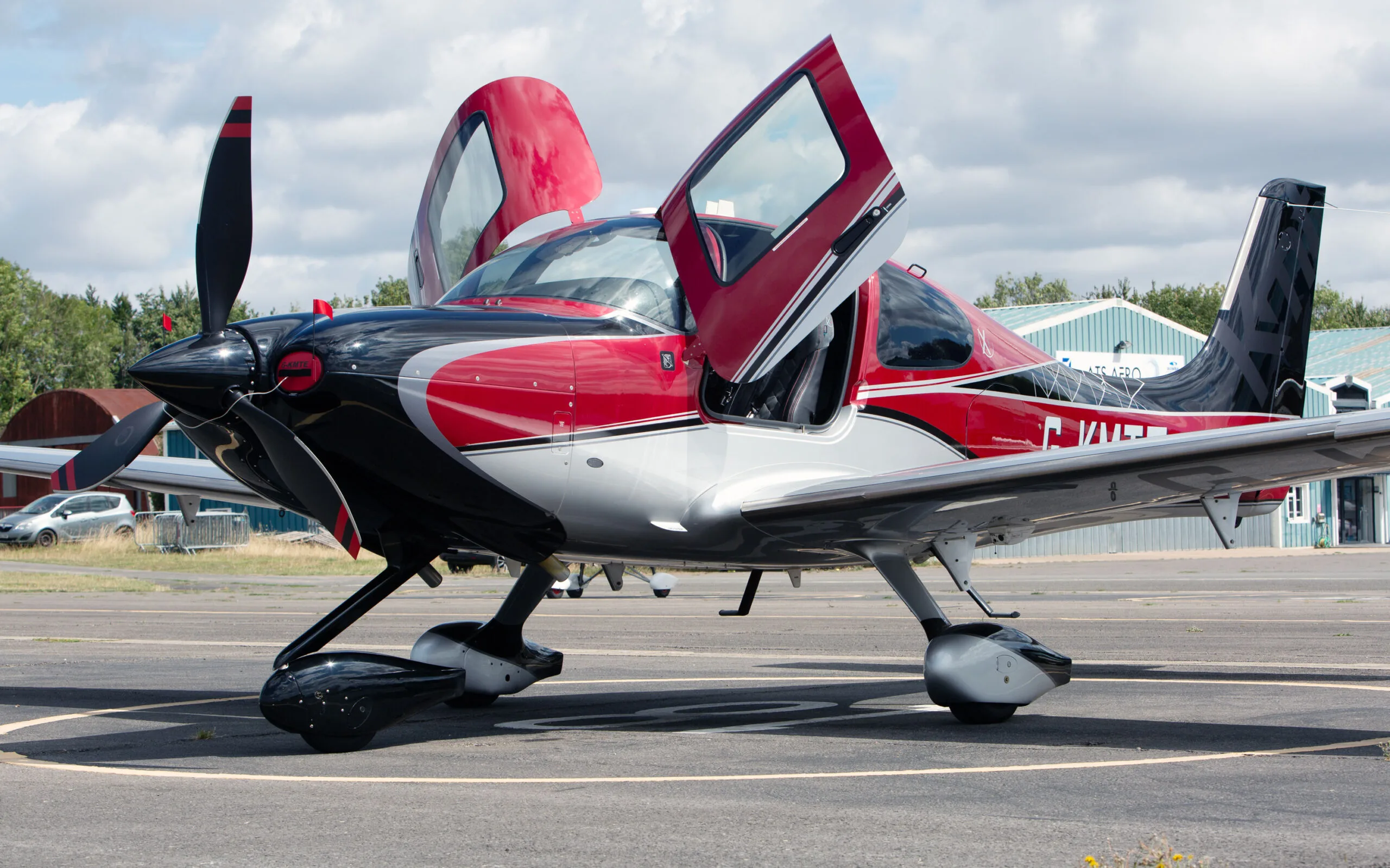 A red, black, and silver Cirrus SR22 aircraft with its gull-wing doors open, parked on an airfield under a cloudy sky.