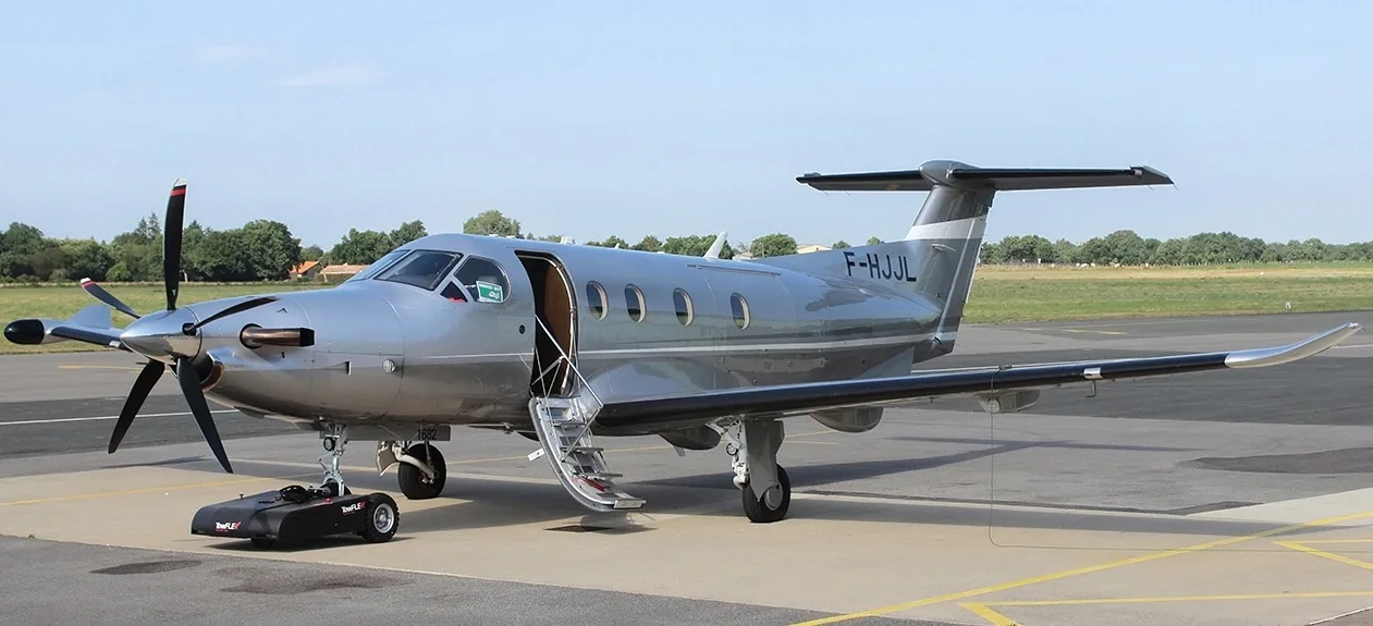 A silver Pilatus PC-12 aircraft with its main door open and stairs extended, parked on an airfield with a small ground tug nearby.
