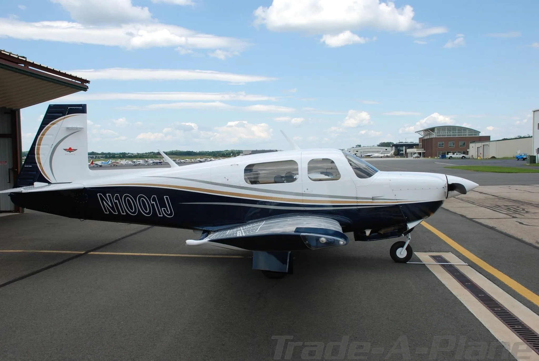 A white and blue Mooney aircraft (N1001J) parked on a tarmac with hangars and other aircraft in the background.