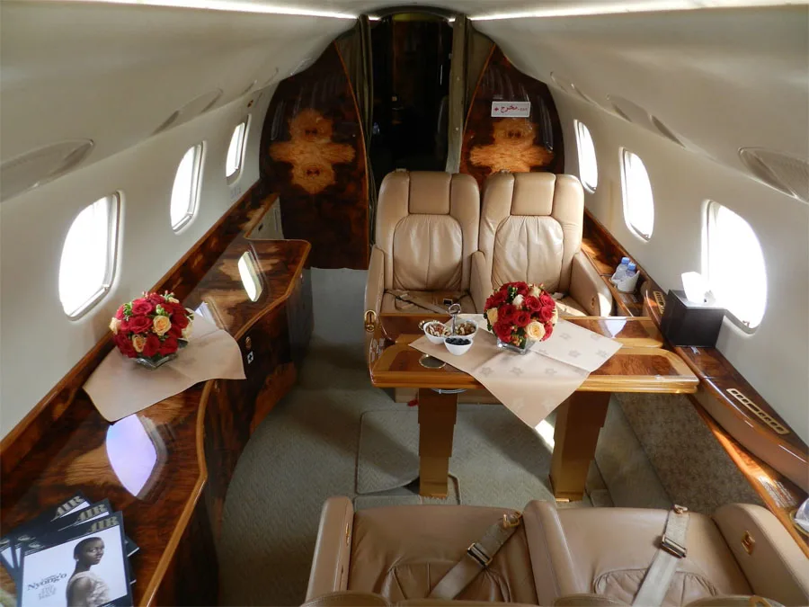 A view inside a private jet cabin shows a dining table, leather seats, and polished wood paneling.