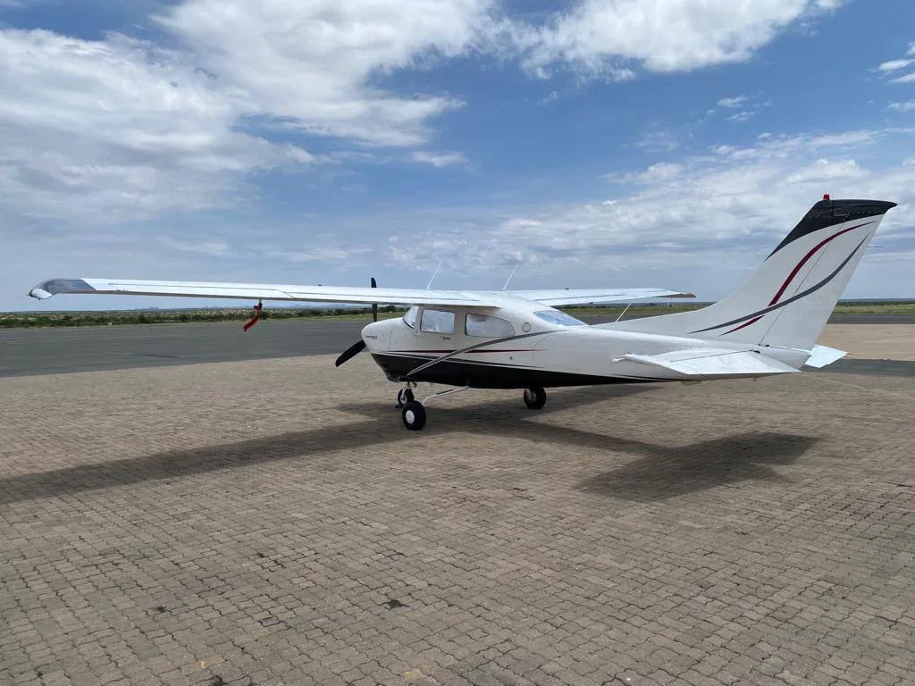 A white single-engine propeller aircraft with black and red accents on the tail, parked on a paved surface under a partly cloudy sky.