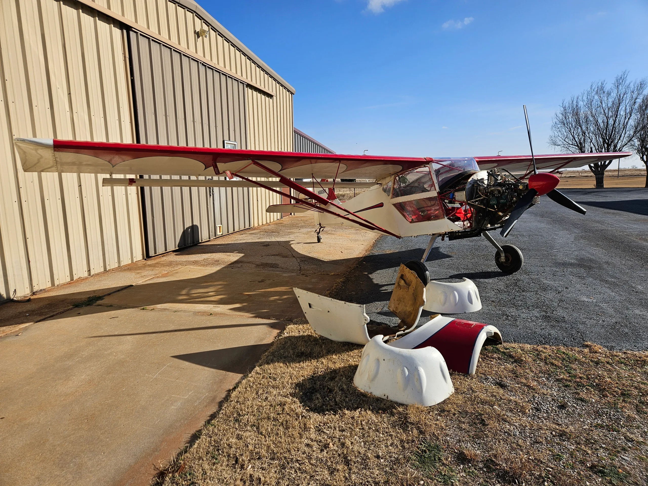 A white and red experimental aircraft with its engine cowling removed, parked on an asphalt apron next to a hangar under a blue sky.