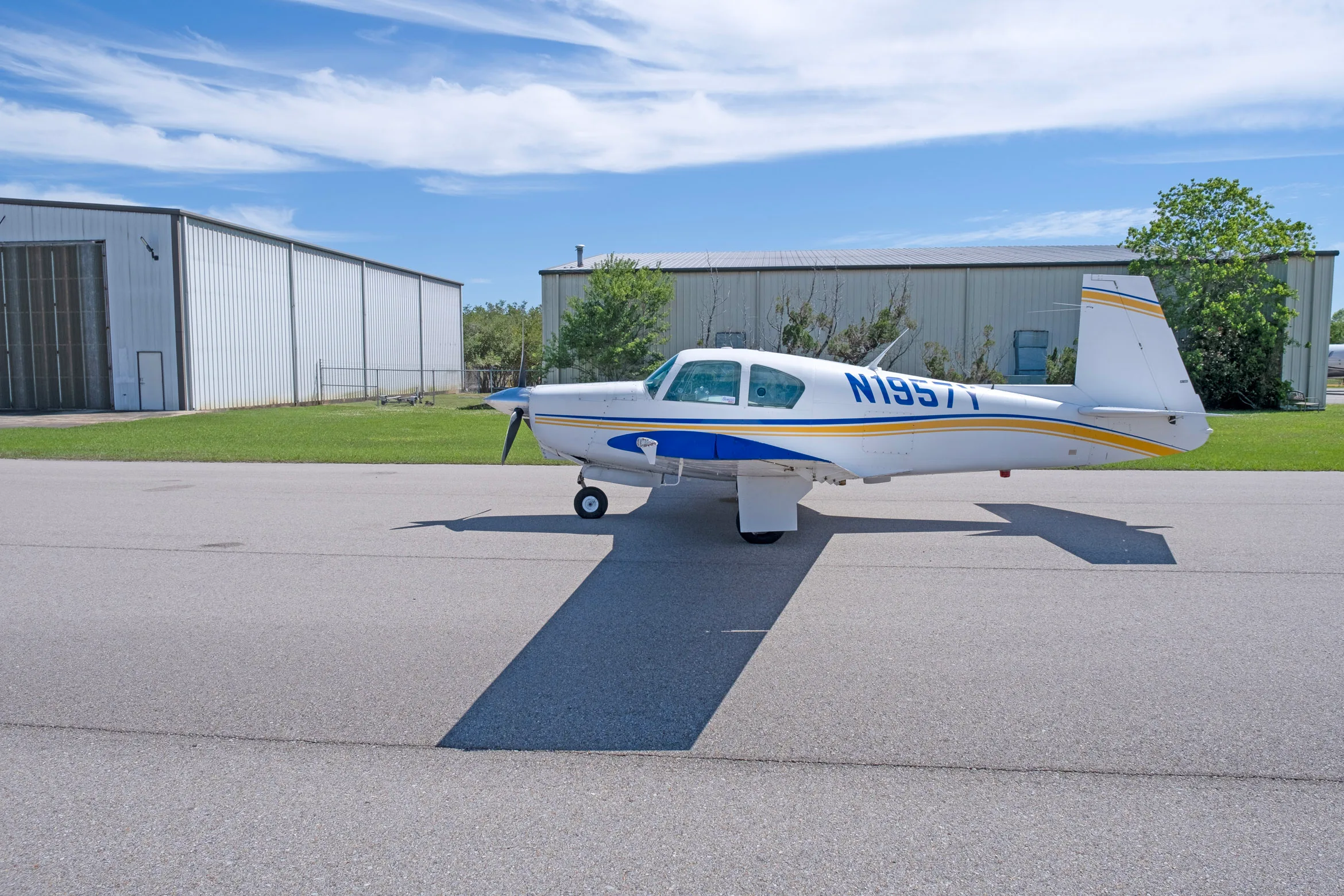 A side view of a white single-engine propeller aircraft with blue and yellow stripes, registration N1957Y, parked on an asphalt apron.