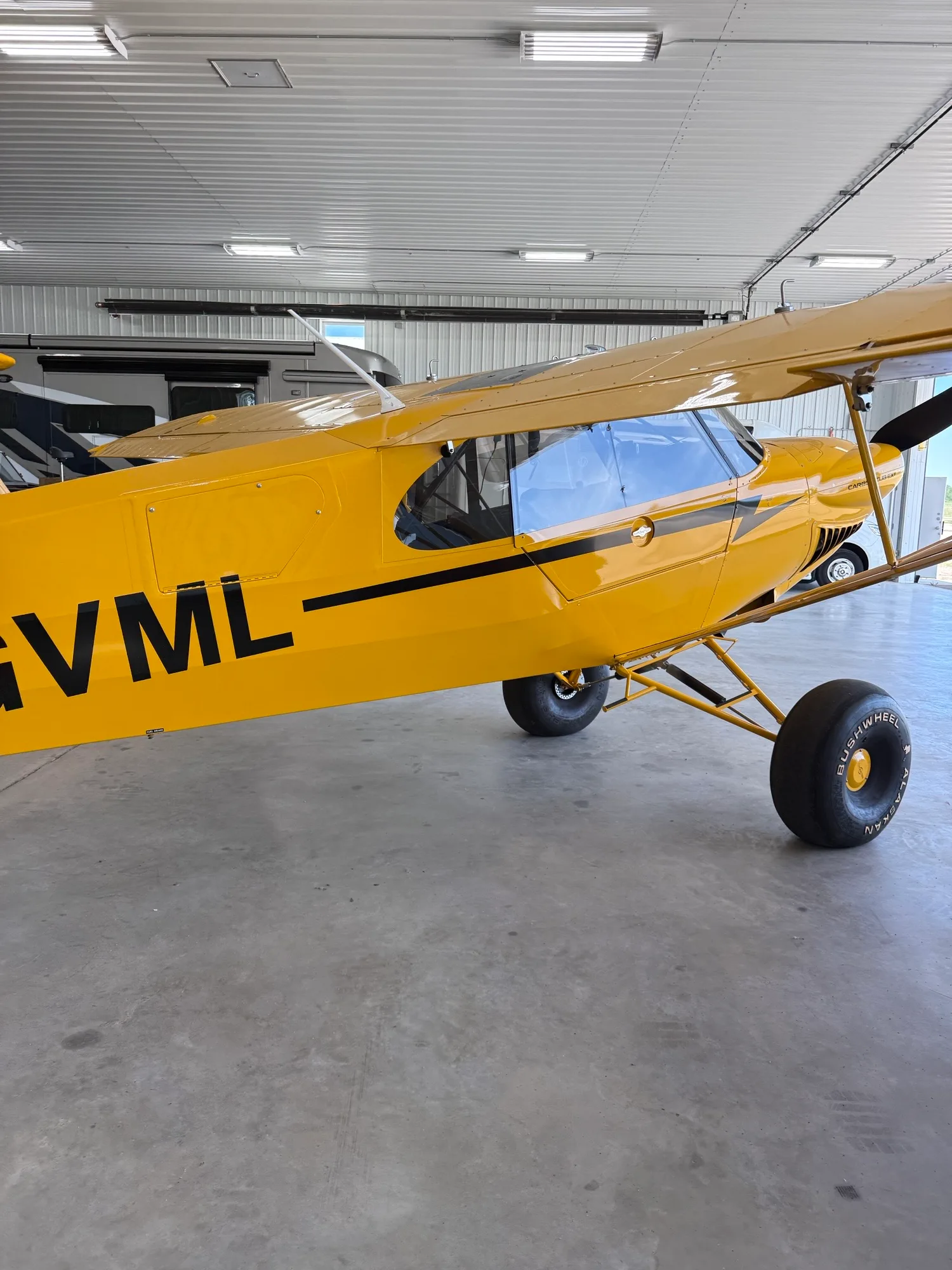 The side view of a yellow CubCrafters Carbon Cub EX-3 with registration C-GVML and large tires, inside a hangar.